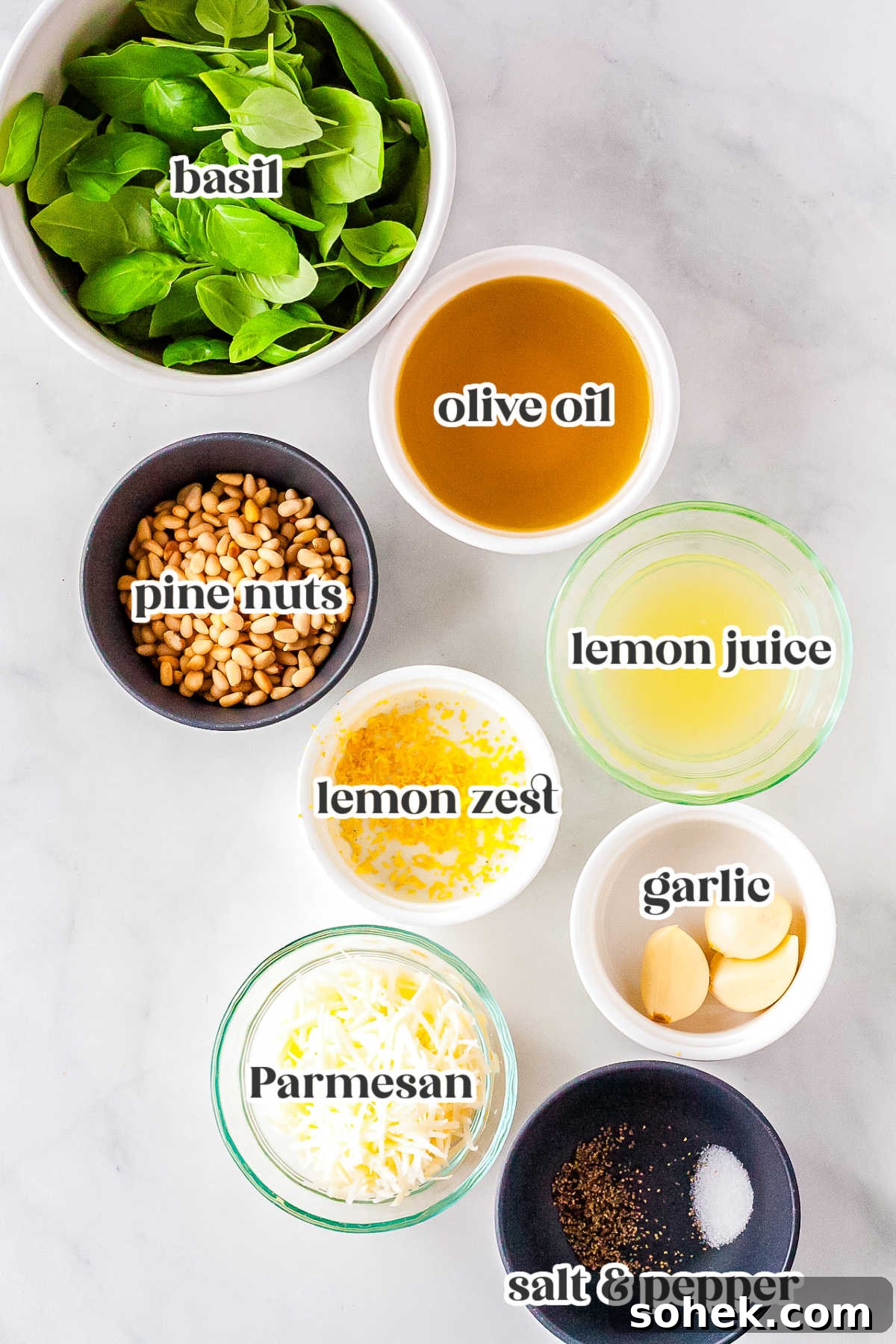 A flat lay of the ingredients needed for the recipe including fresh basil, olive oil, pine nuts, lemon juice, lemon zest, garlic cloves, Parmesan cheese, salt, and black pepper.