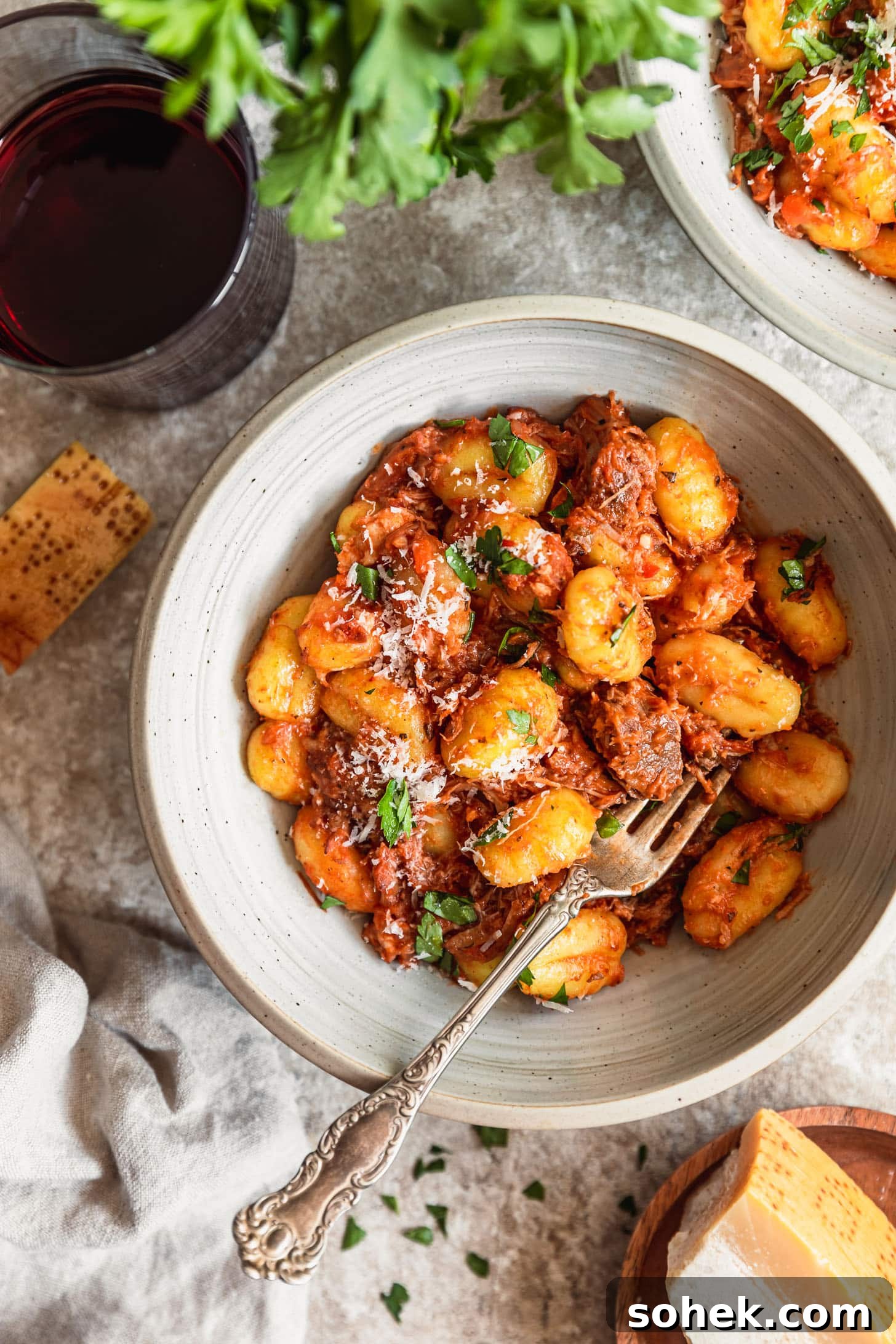 A white bowl with braised pork ragù with gnocchi on a beige counter next to another bowl of pasta, red wine, a wood bowl of parmesan, and parsley.