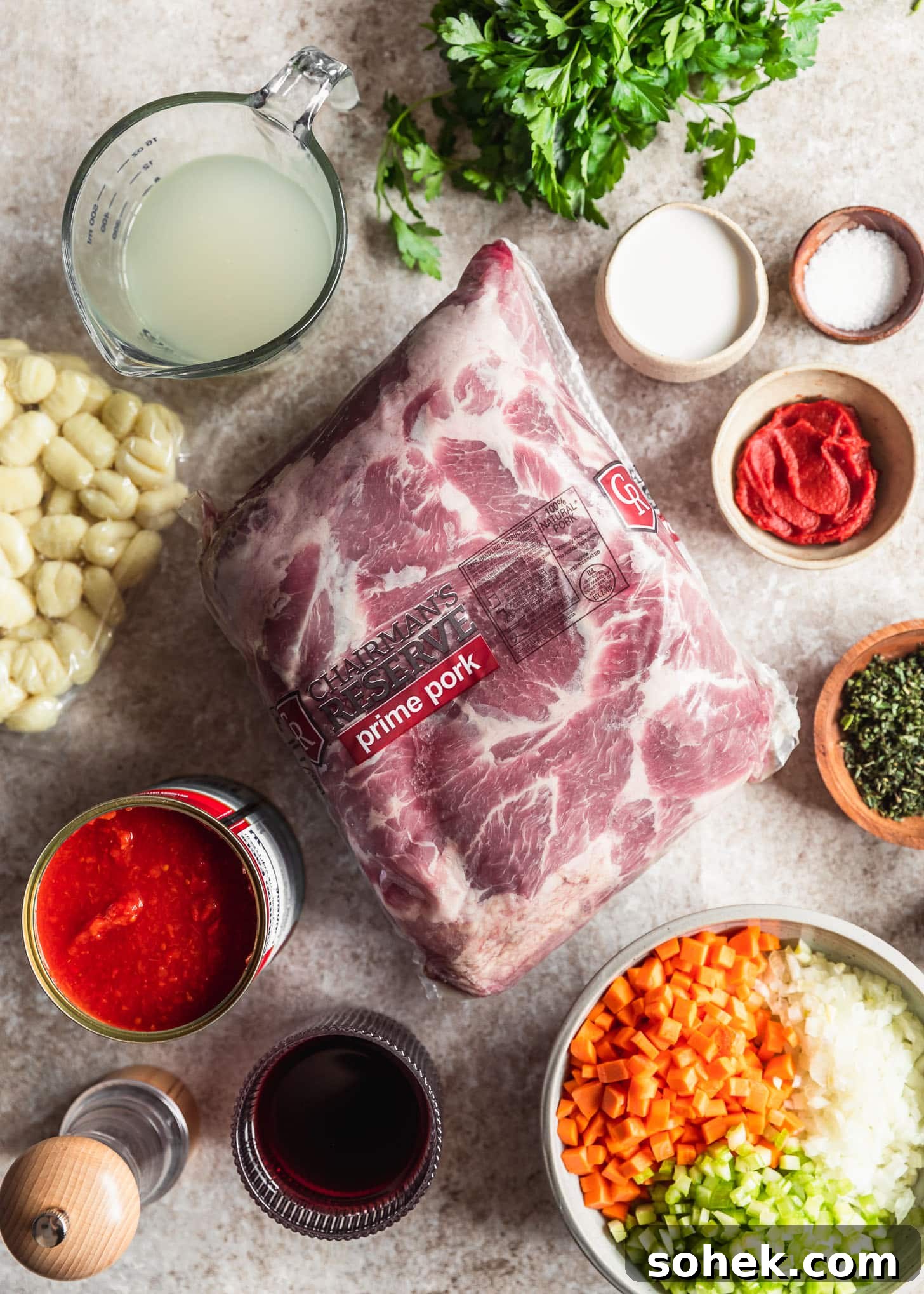 Pork shoulder next to parsley, a can of tomatoes, wine, pasta, and white bowls of carrots, celery, onions, tomato paste, and milk on a tan counter.