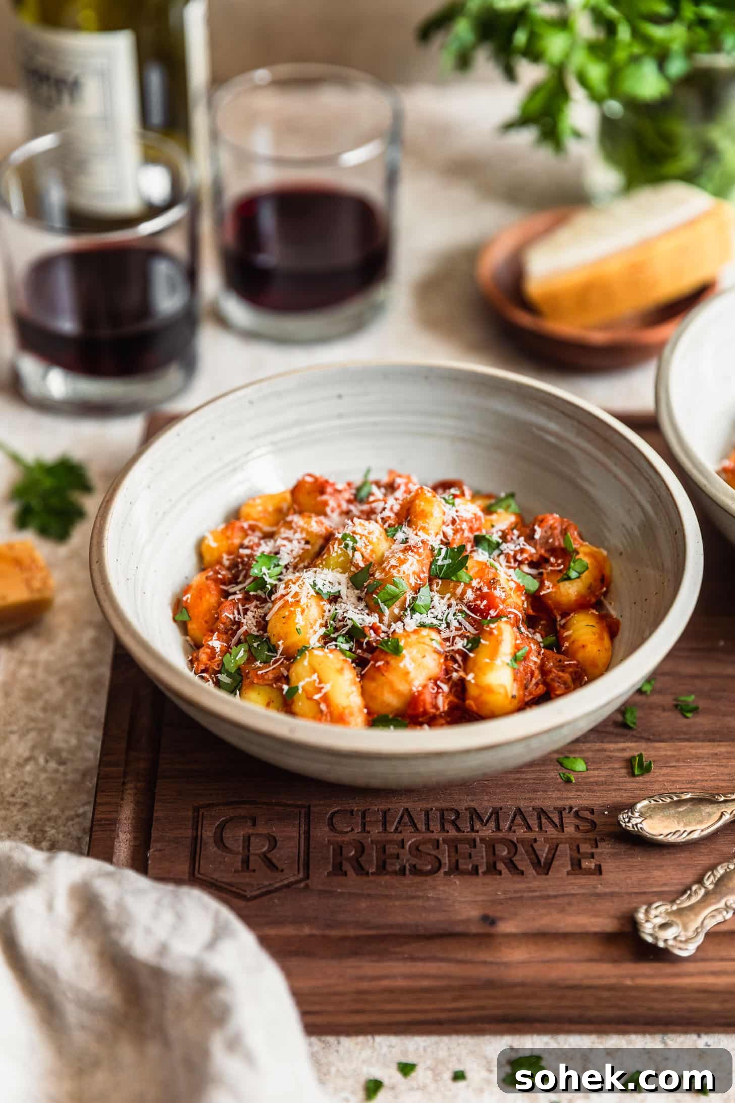 A white bowl with pork ragù with gnocchi on a wood board next to glasses of red wine, a jar of parsley, forks, and a beige linen on a tan counter.