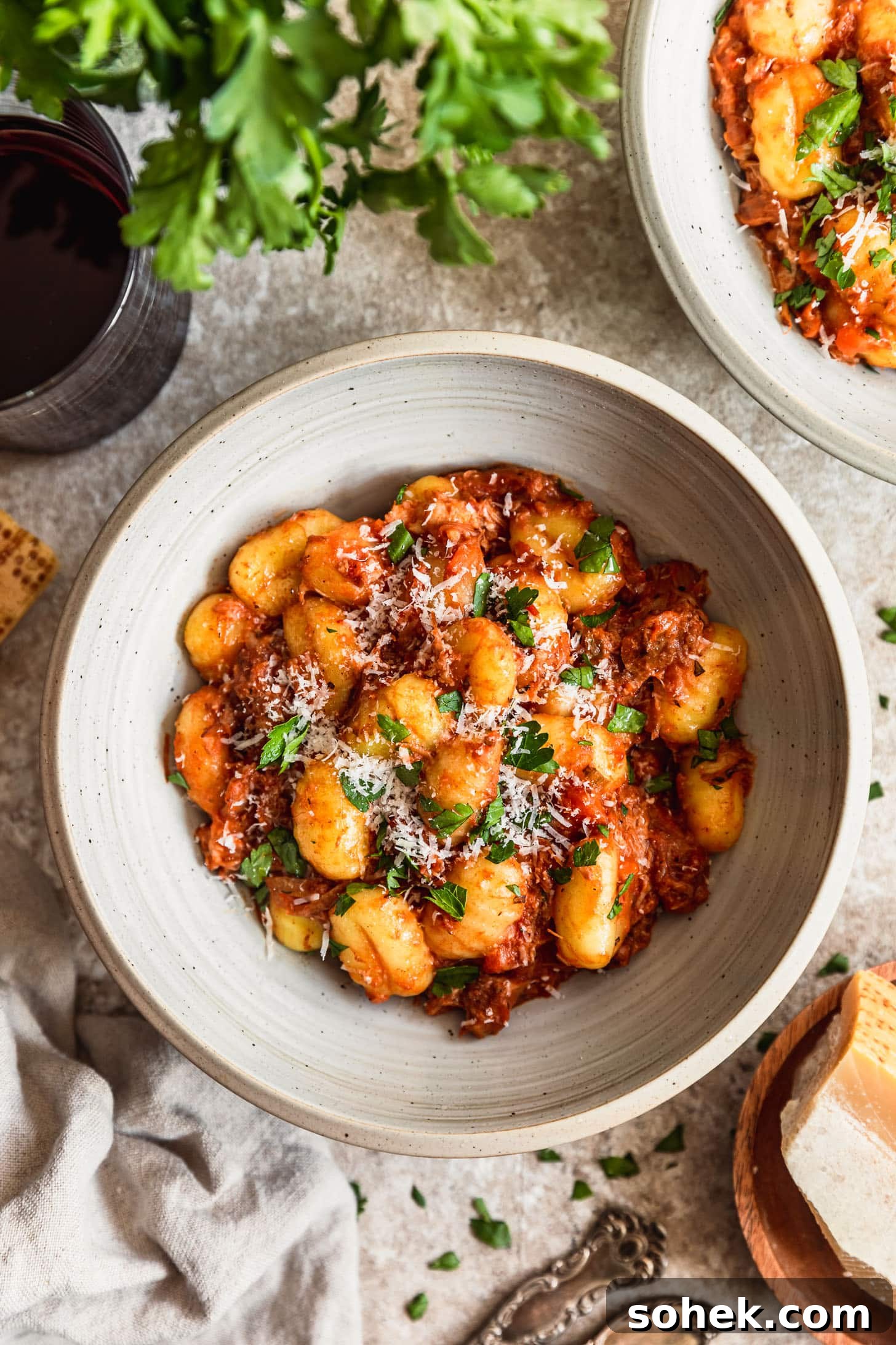 A white bowl of pork ragù with gnocchi on a beige table next to Parmesan, red wine, parsley, and a tan linen.