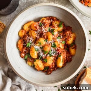 A white bowl of pork ragu with gnocchi on a beige table next to parmesan and red wine.