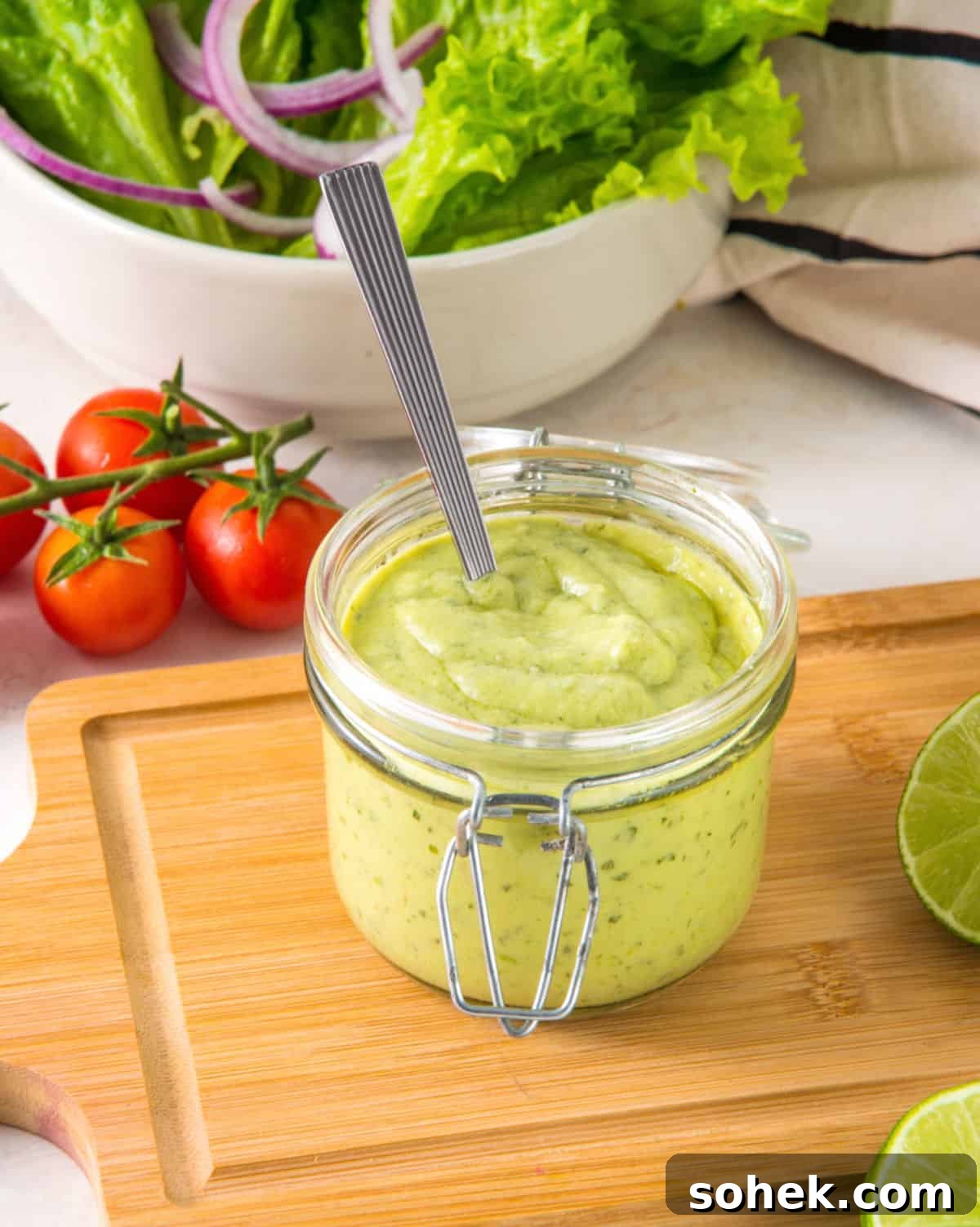 Avocado ranch dressing in a mason jar on a wooden board. Tomatoes, bowl with greens, limes.