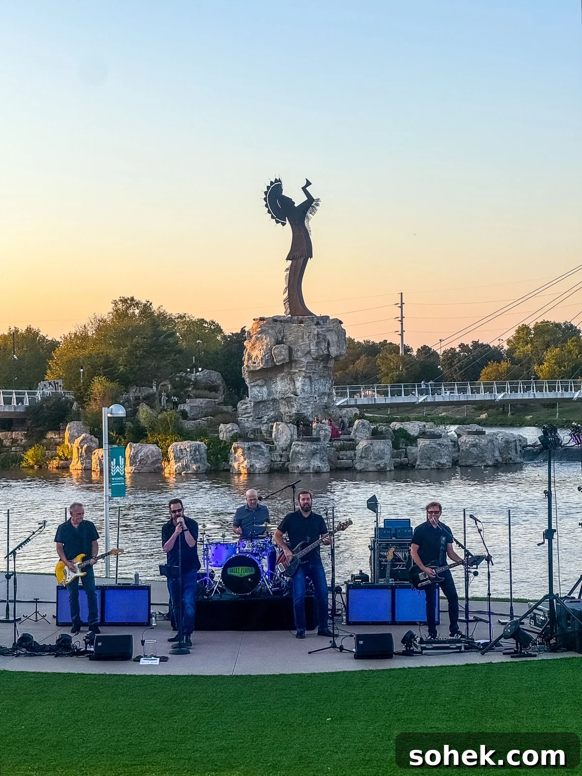 Discover Wichita, Kansas: Must-See Sights and Local Gems 2 A band performs outdoors on a stage in front of a river, with the Keeper of the Plains statue on a rocky pedestal in the background at sunset.