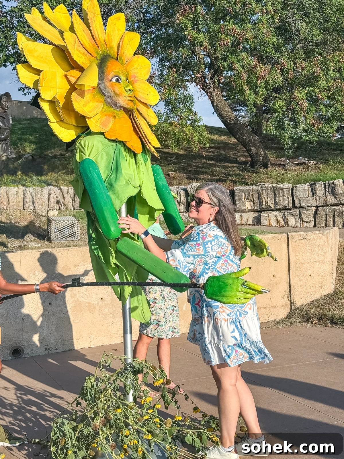Discover Wichita, Kansas: Must-See Sights and Local Gems 11 A woman in a patterned dress interacts with a large person-sized sunflower puppet outdoors on a sunny day.