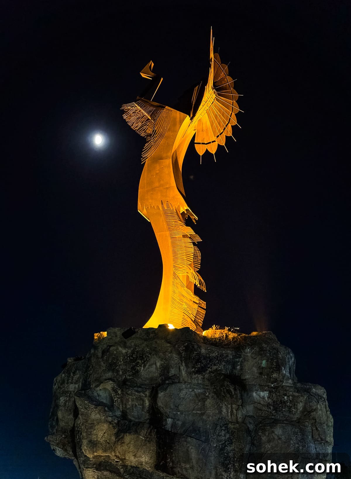 Discover Wichita, Kansas: Must-See Sights and Local Gems 7 A tall, illuminated metal sculpture of a Native American woman stands on a rocky base at night, with the moon visible in the dark sky behind her.
