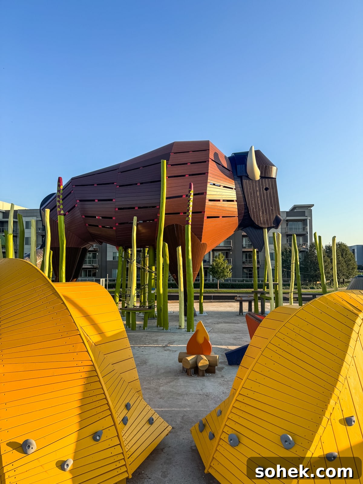 Discover Wichita, Kansas: Must-See Sights and Local Gems 8 A large wooden bison sculpture stands among green poles, with yellow curved play structures and a small campfire sculpture in the foreground. Modern buildings are visible in the background.