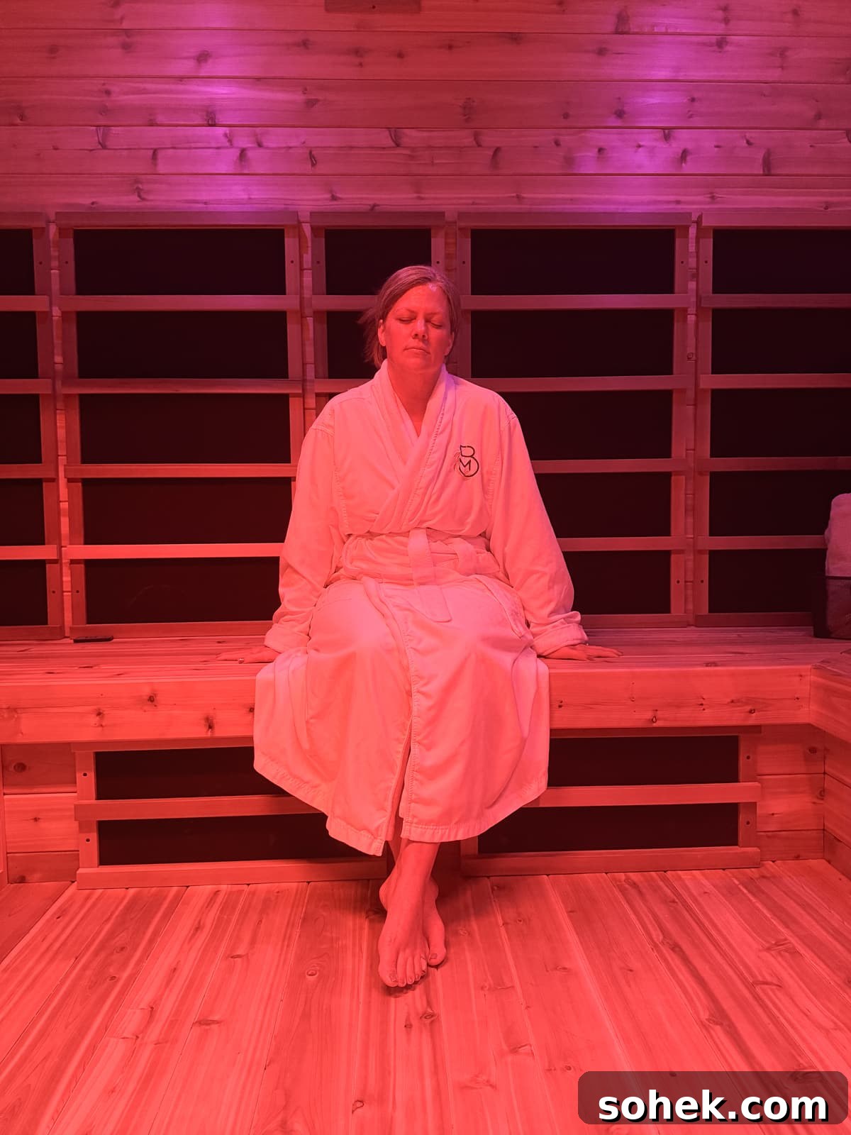 Discover Wichita, Kansas: Must-See Sights and Local Gems 10 A person in a white robe sits on a wooden bench in a sauna room lit with pink light, eyes closed and hands resting on their lap.