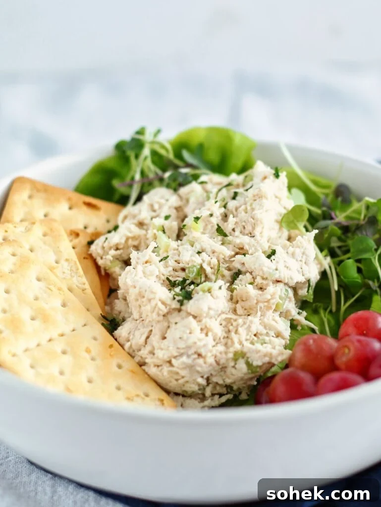 classic chicken salad on a bed of lettuce in a white bowl with grapes and crackers on the side.
