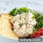 classic chicken salad on a bed of lettuce in a white bowl with grapes and crackers on the side.
