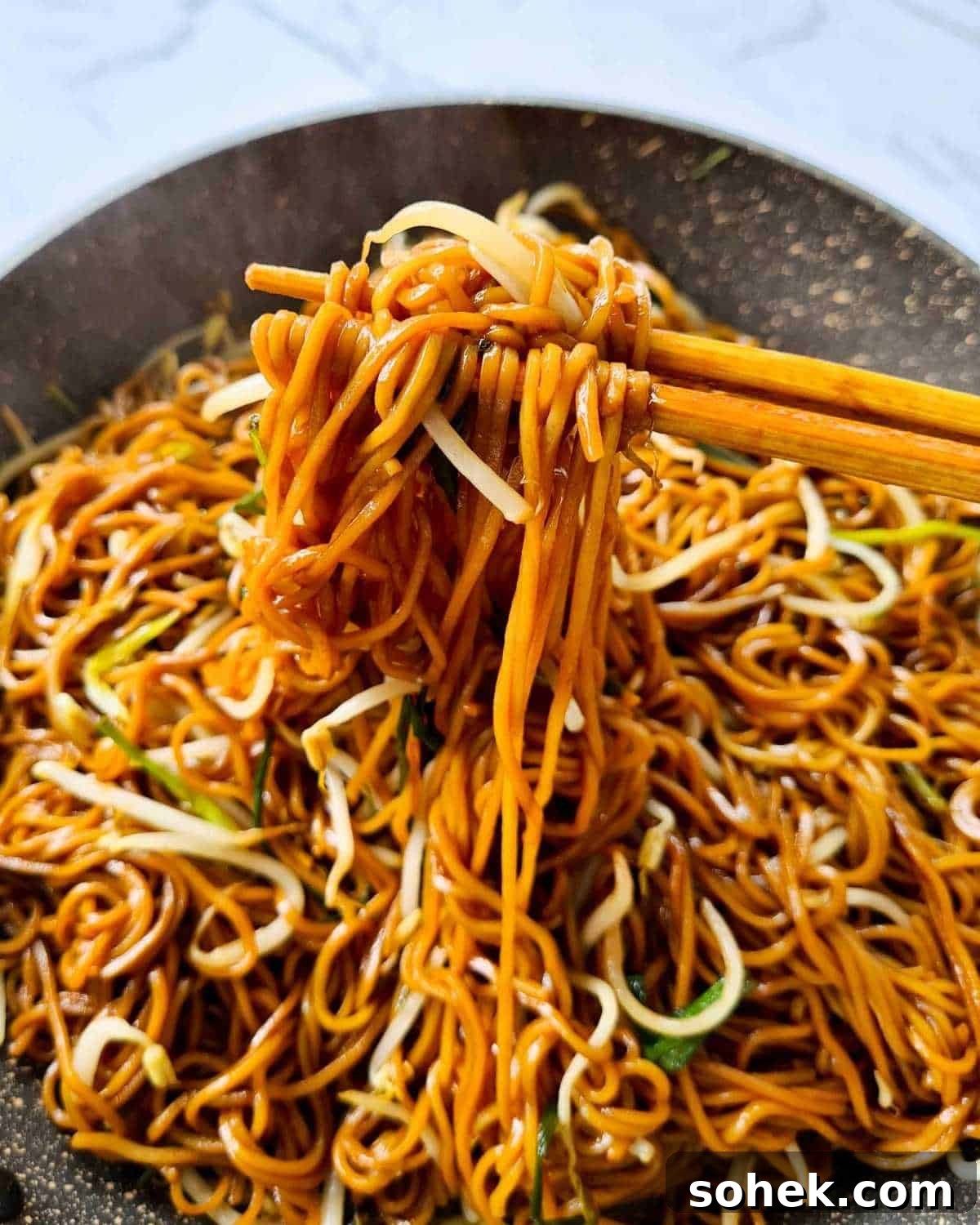 Steaming hot soy sauce noodles being picked up by a pair of chopsticks over a pan full of noodles