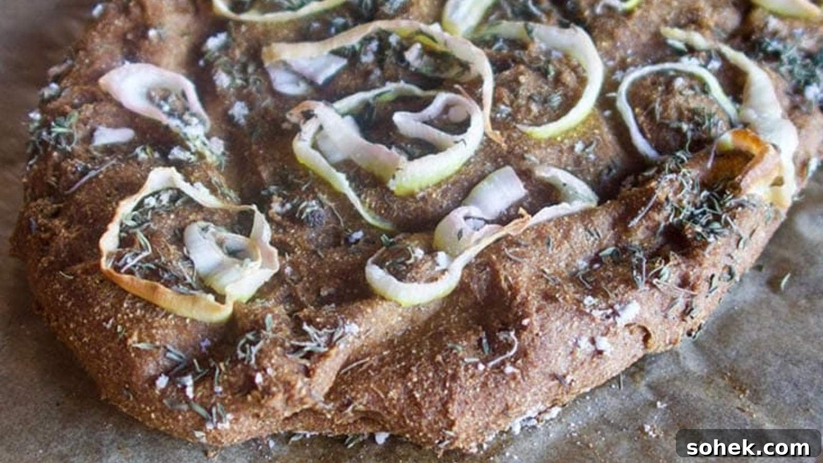 Close-up of a homemade focaccia bread with onions and herbs on a textured surface.