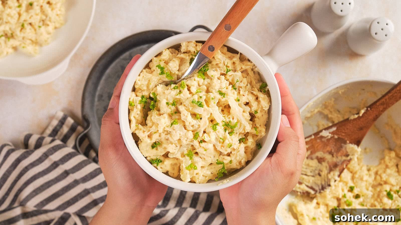 Hands holding a white bowl of macaroni salad garnished with chopped herbs.
