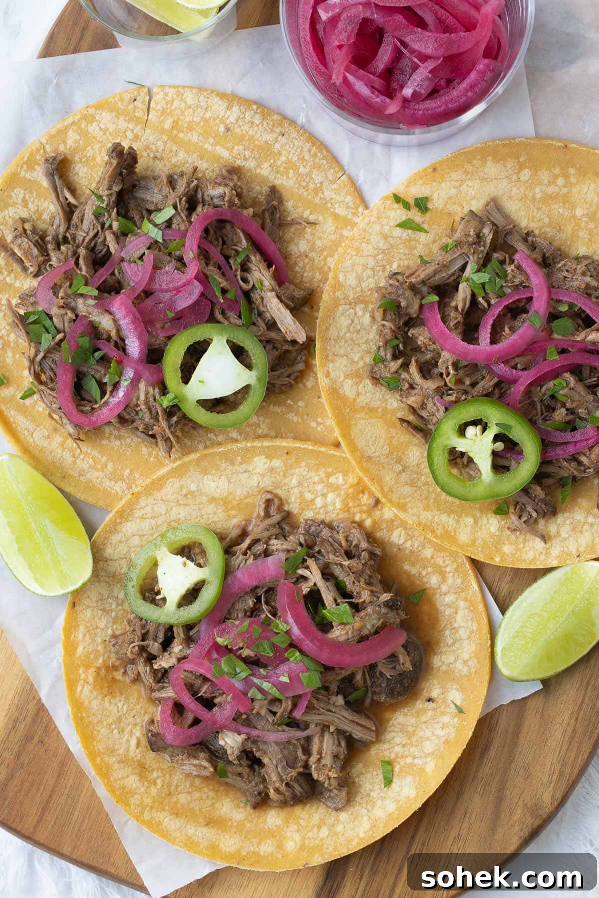 Slow Cooker Shredded Beef Recipe for Sandwiches and Tacos 5 Three shredded beef tacos on golden corn tortillas with pink onions and jalapeno slices.