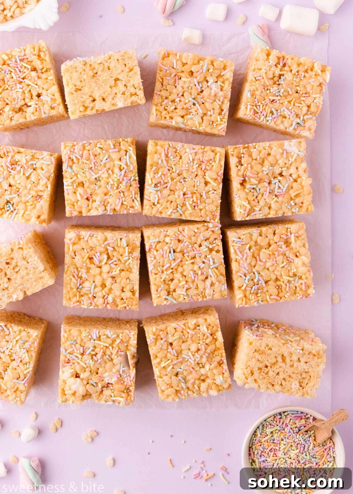 Gluten-Free Rice Krispie Treats Recipe — Crispy & Chewy Bars 11 Squares of gluten free rice krispie treats arranged haphazardly on a sheet of crinkled parchment on a lilac background, with mini marshmallows, rice krispies and sprinkles scattered around.