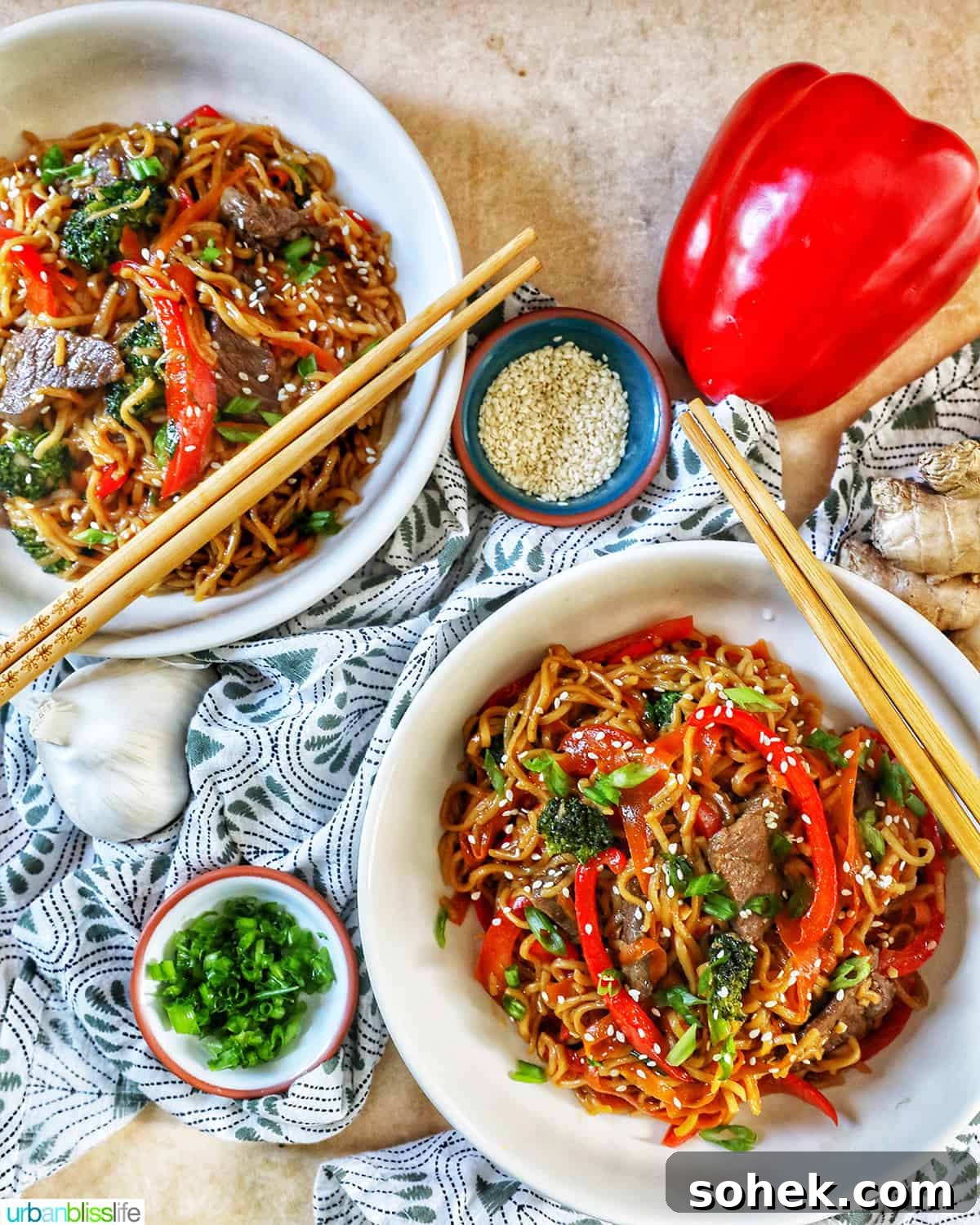 two bowls of sesame garlic noodles with chopsticks, bowls of scallions and sesame seeds, a red bell pepper, all on a green and white napkin and yellow table.
