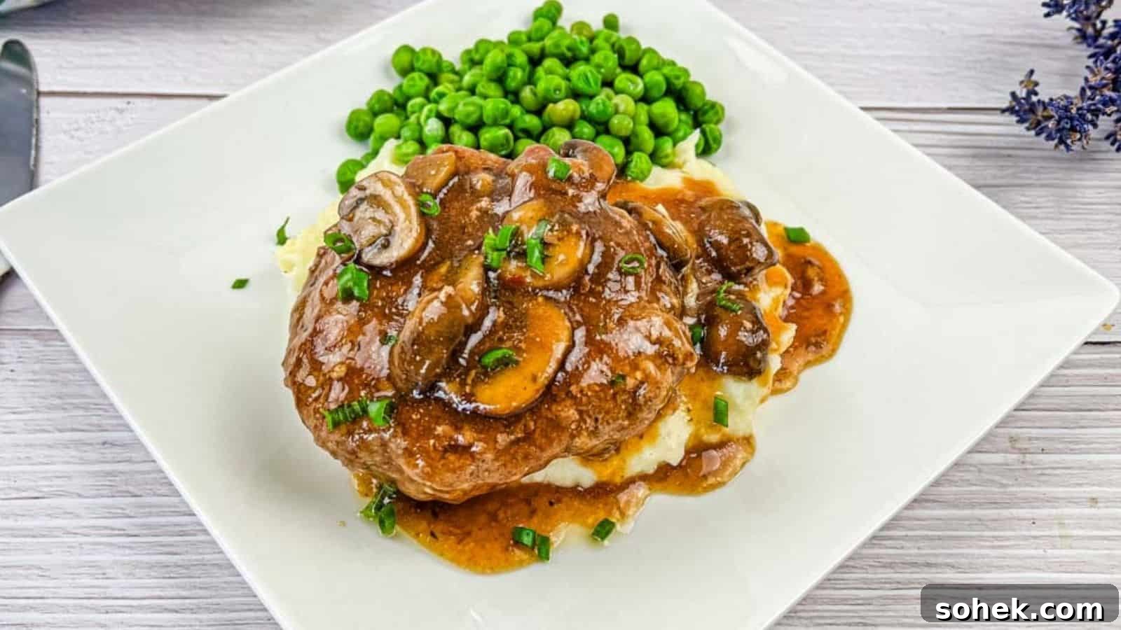 21 Quick Ground Beef Dinners That Will Have Everyone Asking Your Secret 15 A white plate with Salisbury steak topped with mushroom gravy on mashed potatoes, accompanied by a side of green peas.