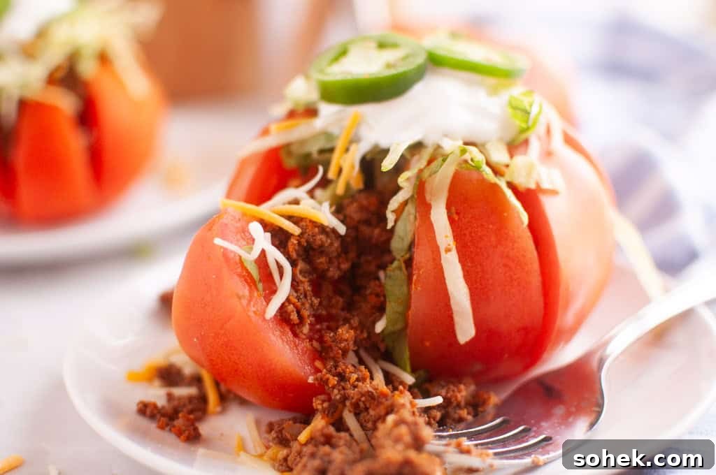 21 Quick Ground Beef Dinners That Will Have Everyone Asking Your Secret 8 A sliced tomato stuffed with seasoned ground meat, shredded cheese, lettuce, sour cream, and jalapeño slices is served on a white plate with a fork.