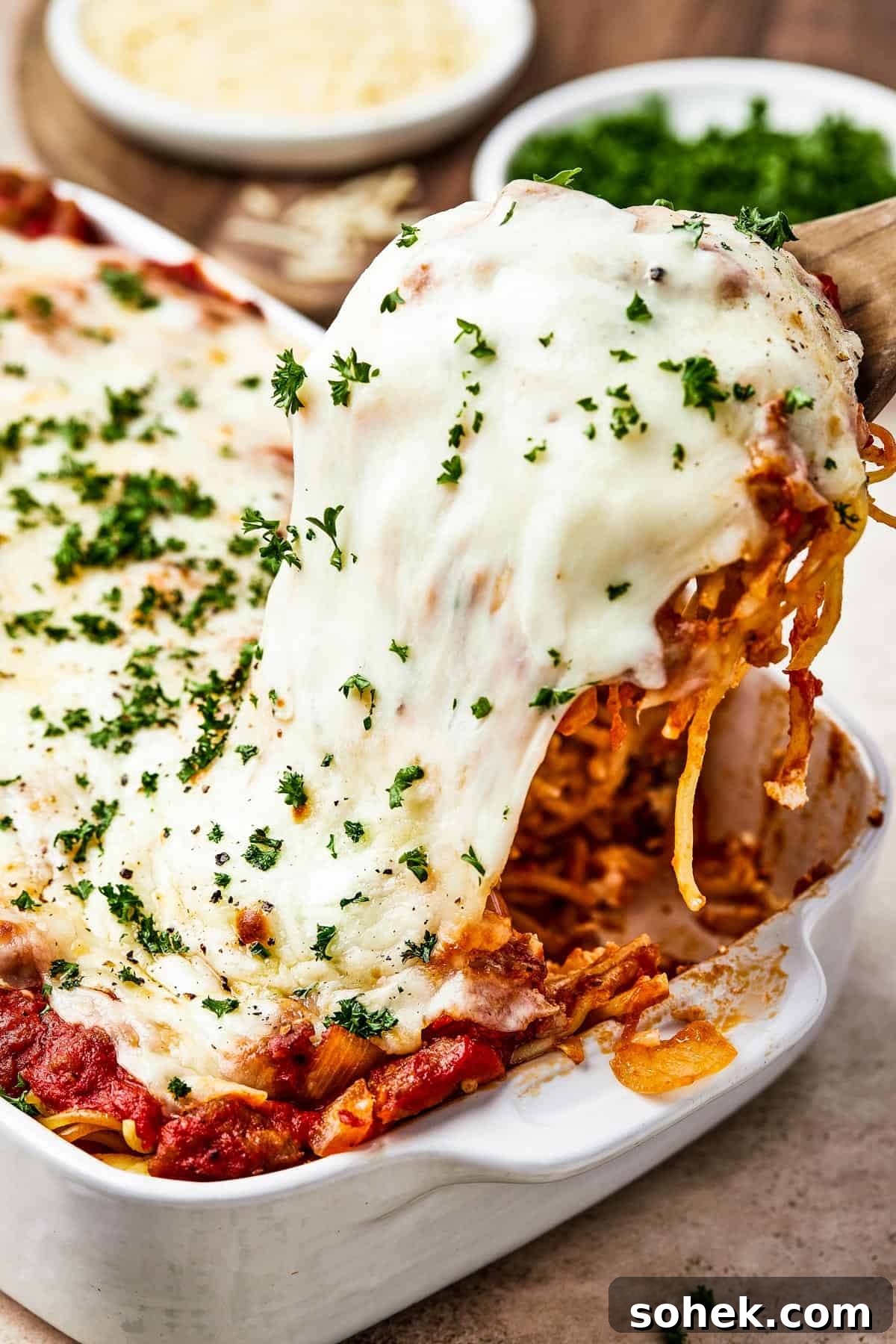 A portion of spaghetti bake being lifted from a casserole dish, with the cheese topping stretching.