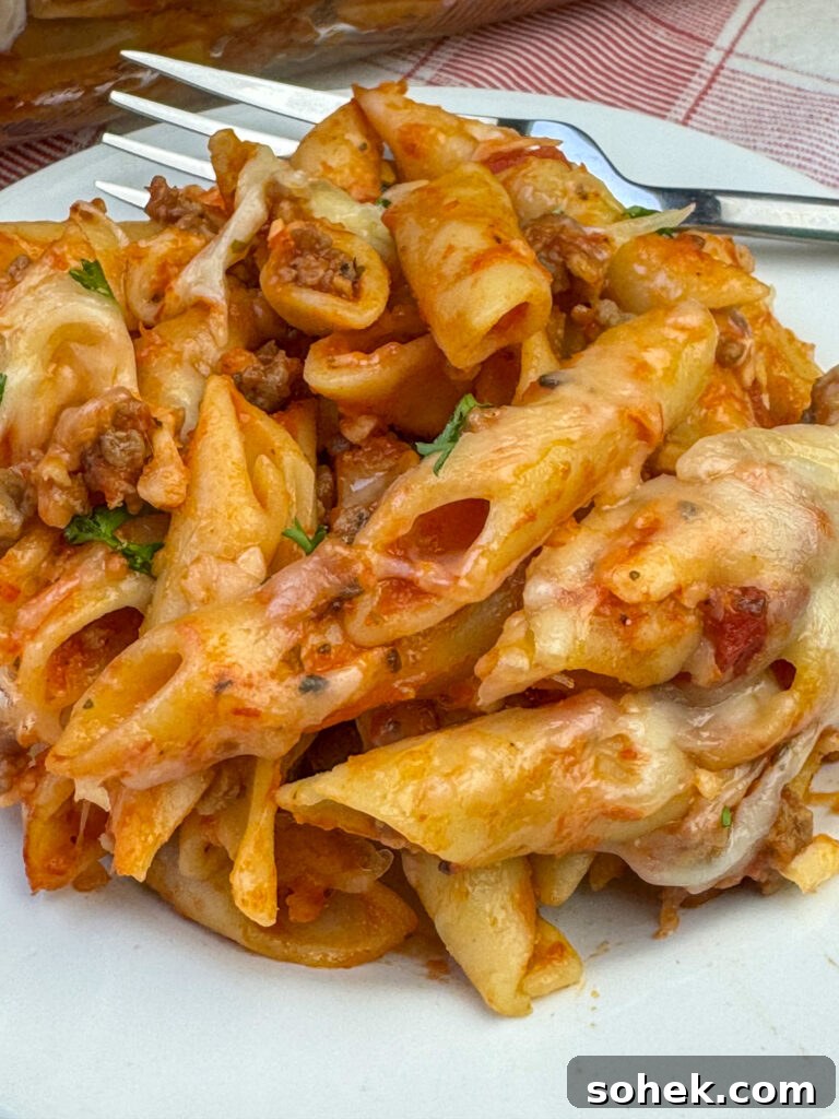 Close up pieces of tender mostaccioli coated in pasta sauce and mozzarella cheese with brown bits of meat in each bite and a fork resting on the back of the plate.
