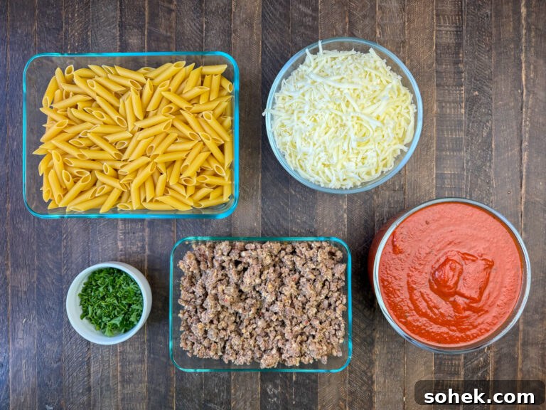 Ingredients to make Mostaccioli are pasta, shredded mozzarella cheese, parsley, Italian sausage and red sauce on a brown counter top.