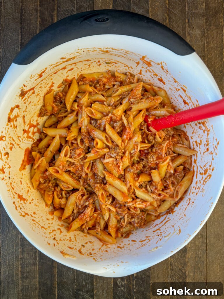 Large mixing bowl with a spoon stirring noodle into a meat, sauce and cheese mixture.
