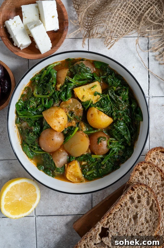 Spinach and potato stew in a white bowl, surrounded by feta, olives and bread.