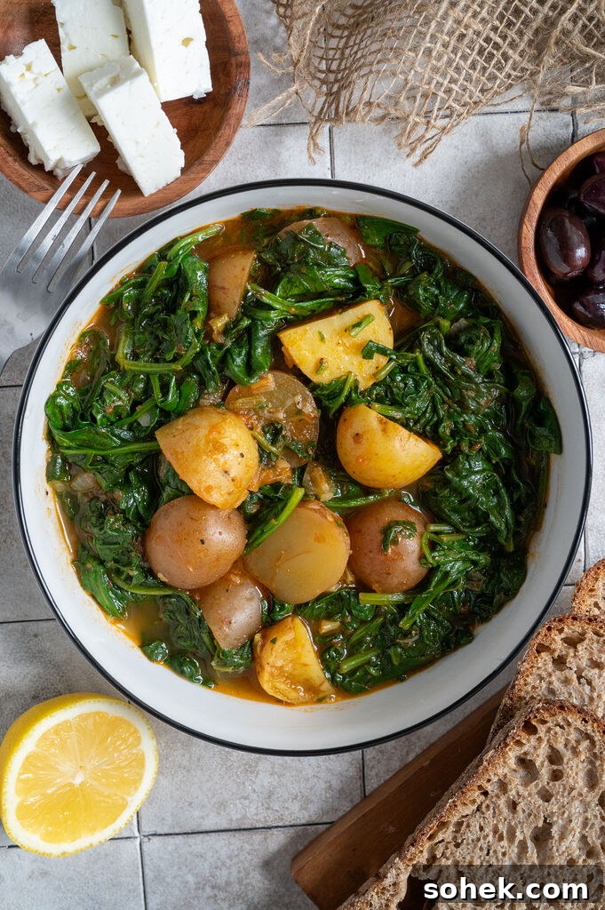 Spinach and potato stew in a white bowl, surrounded by feta, olives and bread.