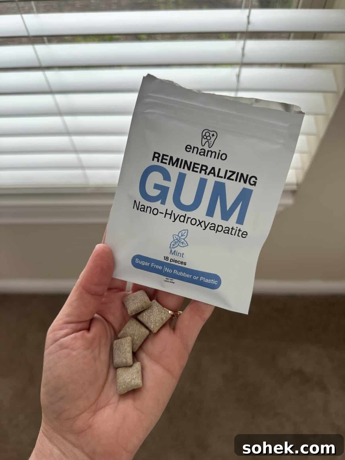 A hand holding several square pieces of beige chewing gum in front of a white package labeled “enamio Remineralizing Gum – Nano-Hydroxyapatite, Mint, 18 pieces, Sugar Free, No Rubber or Plastic.”