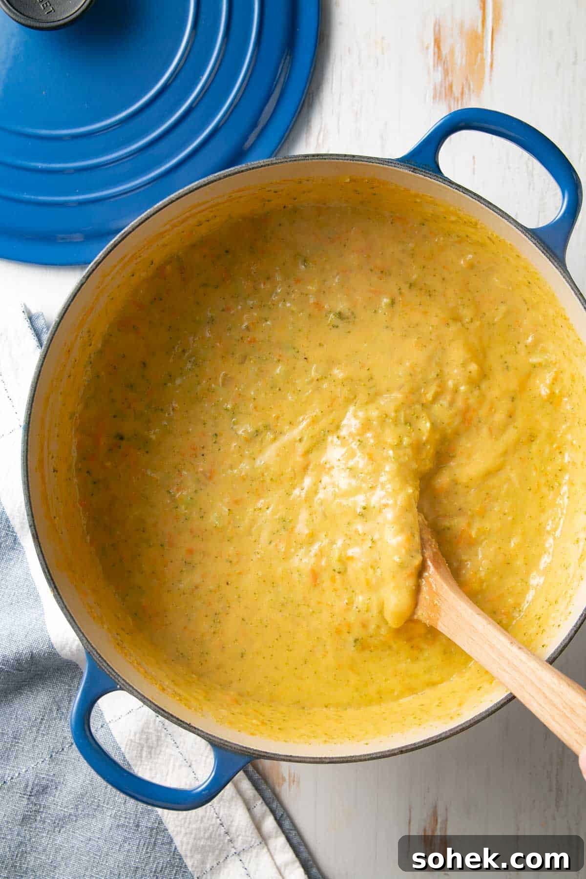 A big blue pot filled with broccoli cheddar soup stirred with wooden spoon.