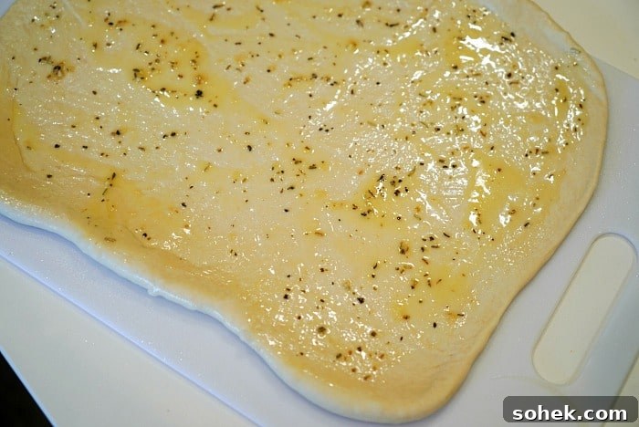 A sheet of uncooked thawed from frozen pizza dough is evenly spread with olive oil and sprinkled with herbs, positioned on a white cutting board.