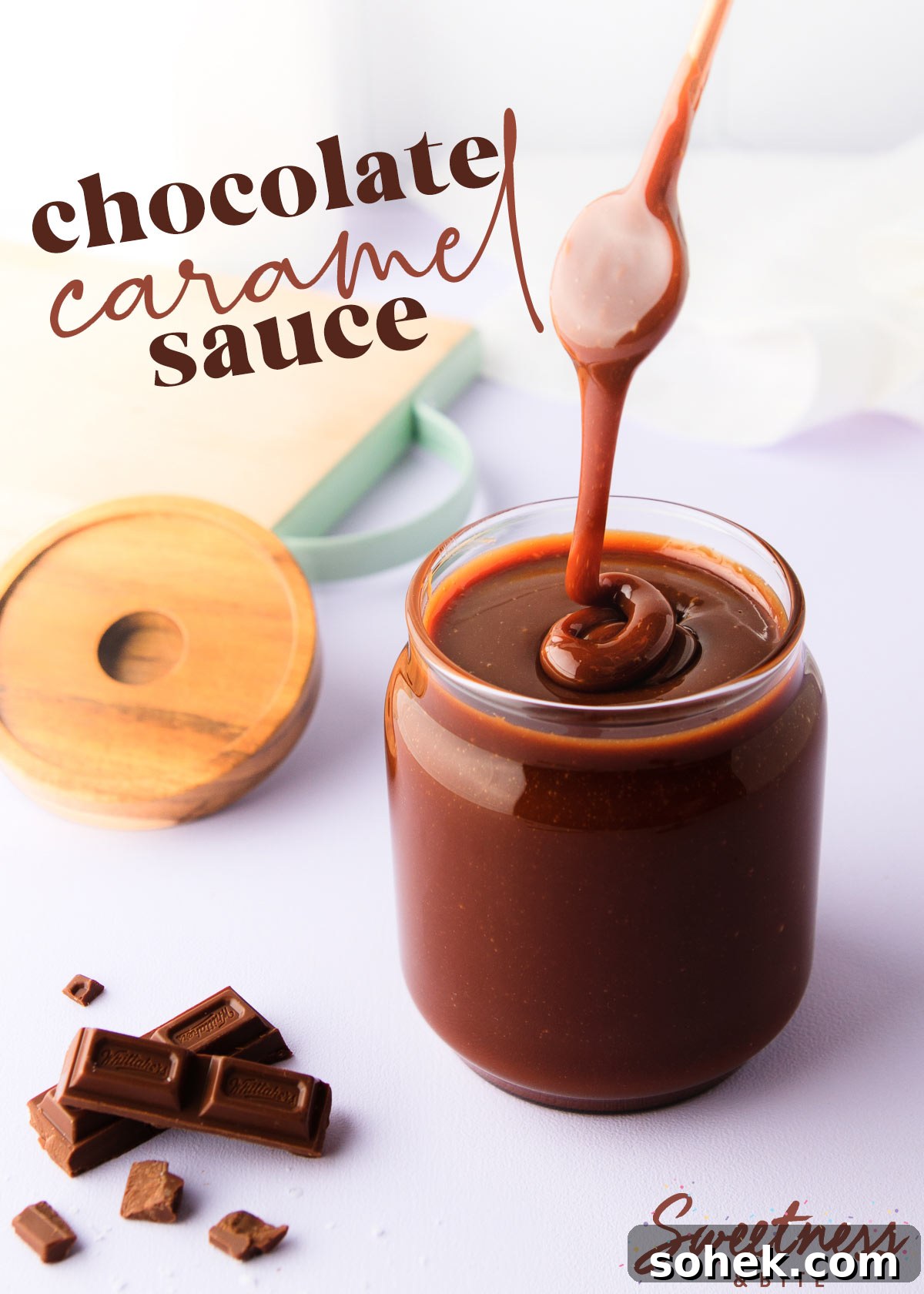 A jar of chocolate caramel sauce on a pale purple background, with a spoon drizzling the thick sauce back into the jar, and some chopped chocolate next to the jar.